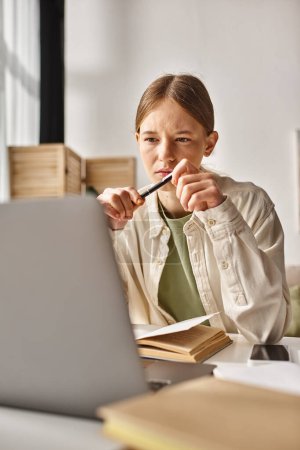 Téléchargez les photos : Adolescent fille tenant stylo et regardant ordinateur portable tout en faisant ses devoirs près de livre et téléphone sur le bureau - en image libre de droit