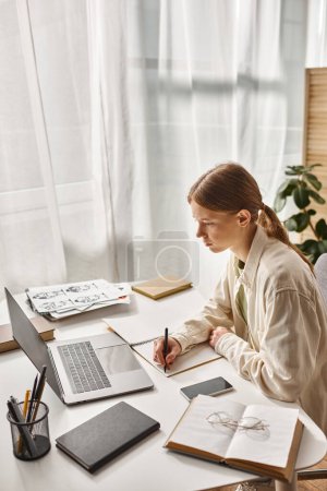 Photo for Teenage girl writing in notebook and looking at laptop while doing her homework, online learning - Royalty Free Image