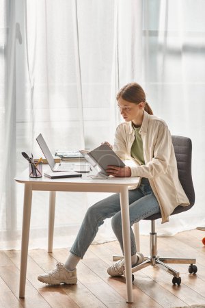 adolescent fille lecture livre tandis que assis près ordinateur portable et papeterie sur le bureau, éducation en ligne