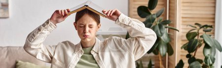 Photo for Overwhelmed teenage girl with a book on her head sitting in front of a laptop at home, banner - Royalty Free Image