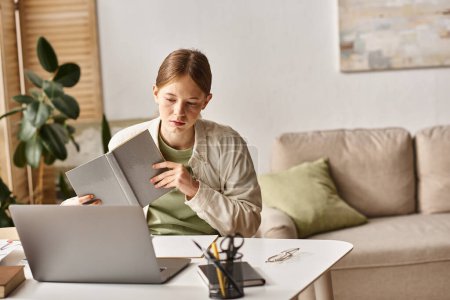geschäftiges Teenager-Mädchen, das sein Lernbuch in der Hand hält und zu Hause vor einem Laptop sitzt, E-Learning