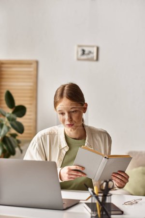 umtriebige Teenager-Studentin hält ihr Lernbuch in der Hand und sitzt zu Hause vor einem Laptop, E-Learning
