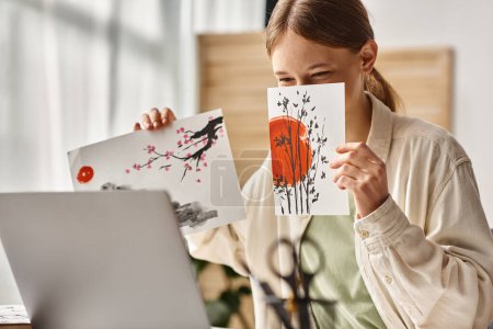 Photo for Teenager girl showing her artwork while covering face and looking at her laptop, art class online - Royalty Free Image