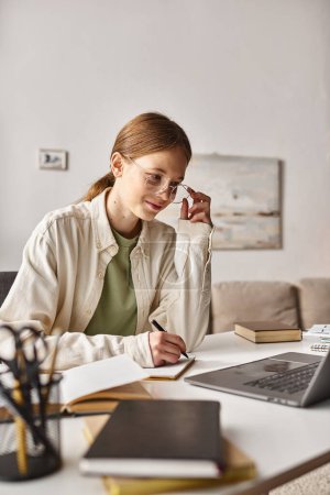 Teenager mit Brille hält Stift in der Hand und schreibt während des Online-Unterrichts am Laptop Notizen