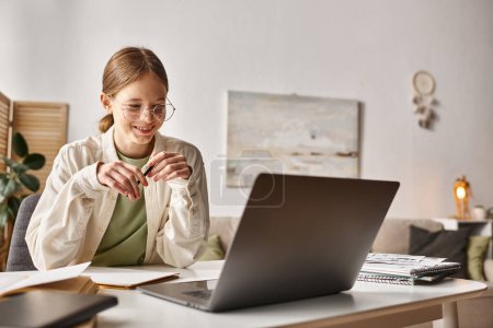 Téléchargez les photos : Heureuse adolescente dans des lunettes tenant stylo et écriture pendant les cours en ligne sur ordinateur portable, prendre des notes - en image libre de droit