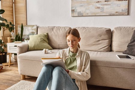 Chica adolescente pensativa leyendo su cuaderno cerca del sofá con auriculares y tableta digital cerca