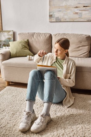 Pensive teenage girl reading her notebook and sitting near the couch with headphones nearby