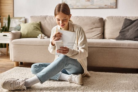 happy teenager girl holding her notebook and smartphone and sitting near the couch with headphones