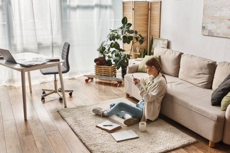 Téléchargez les photos : Adolescent heureux fille en utilisant son smartphone tout en étant assis sur le tapis près du canapé dans le salon - en image libre de droit