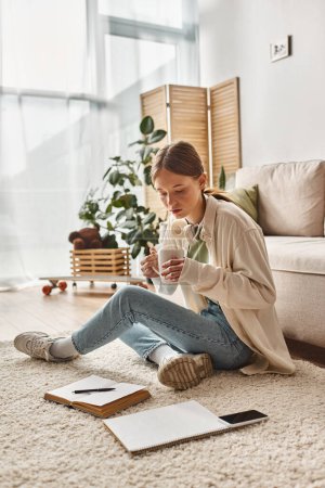 Teen girl enjoys a coffee break at home and looking at smartphone and notebook on carpet