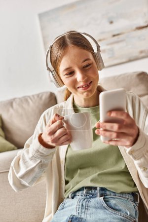 carefree teenager girl in wireless headphones using smartphone while holding cup of tea at home
