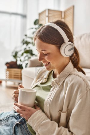 carefree teenager girl in her wireless headphones listening music and holding cup of tea at home