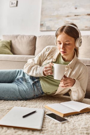 Relaxed teenage girl enjoying music with headphones and holding a mug near notebooks on carpet