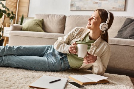 Relaxed gen z girl enjoying music with headphones and holding a mug near notebooks on carpet