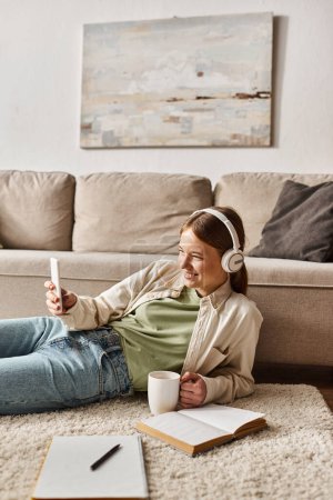 Relaxed gen z girl enjoying music in headphones and taking selfie while holding a mug near notebooks