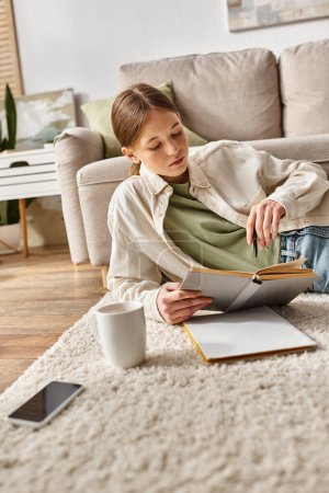 Adolescente niña leyendo libro mientras hace la tarea entre el dispositivo, y la taza de té en la alfombra, generación z