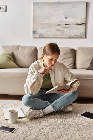 Teenagermädchen liest Buch, während sie Hausaufgaben zwischen Geräten und einer Tasse Tee auf dem Teppich macht, Generation z