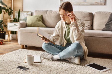 smart teen girl reading book while doing homework among devices and cup of tea, generation z