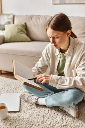 Smart Teenager-Mädchen liest Buch, während sie ihre Hausaufgaben macht und sitzt auf Teppich in der Nähe einer Tasse Tee