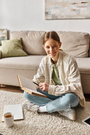 fröhliches Teenager-Mädchen liest Buch, während sie ihre Hausaufgaben macht und auf Teppich in der Nähe einer Tasse Tee sitzt