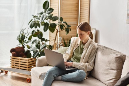 Téléchargez les photos : Adolescente axée sur e-learning à l'aide de son ordinateur portable et assis sur un canapé confortable à la maison - en image libre de droit