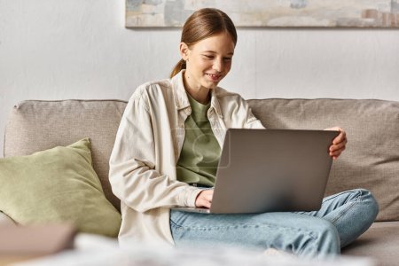 lächelndes Teenie-Mädchen, das sich auf E-Learning mit seinem Laptop konzentriert und zu Hause auf einem bequemen Sofa sitzt