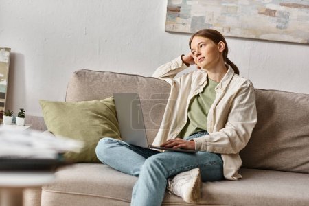 Téléchargez les photos : Adolescente rêveuse assis avec son ordinateur portable sur un canapé confortable à la maison, concept d'apprentissage à distance - en image libre de droit