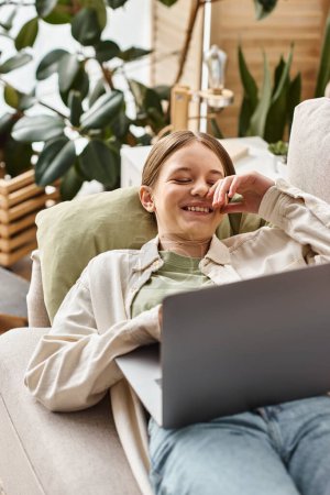 Téléchargez les photos : Heureuse adolescente en utilisant son ordinateur portable et couché sur un canapé confortable à la maison, enseignement à distance - en image libre de droit