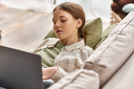 Téléchargez les photos : Adolescente axée sur e-learning à l'aide de son ordinateur portable et couché sur un canapé confortable à la maison - en image libre de droit