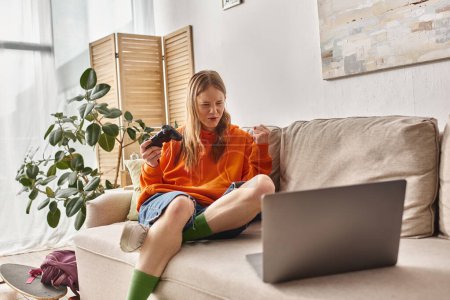 emotional teen girl with joystick and laptop winning game and sitting on sofa at home, weekend vibes