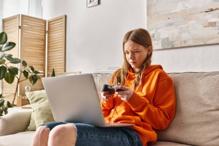 Téléchargez les photos : Adolescent fille avec joystick et ordinateur portable jouer jeu et assis sur canapé à la maison, week-end vibes - en image libre de droit
