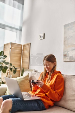 Jovencita alegre jugando con un joystick y un ordenador portátil mientras está sentado en el sofá en casa, vibraciones de fin de semana