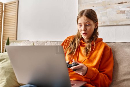 Téléchargez les photos : Jeu adolescent étonnant fille avec joystick et ordinateur portable tout en étant assis sur le canapé à la maison, vibes week-end - en image libre de droit