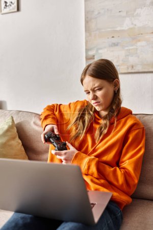 Téléchargez les photos : Jeu adolescent mécontent avec joystick et ordinateur portable tout en étant assis sur le canapé à la maison, divertissement - en image libre de droit