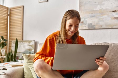 Téléchargez les photos : Heureux adolescent fille avec un ordinateur portable profiter de son temps et assis sur le canapé dans le salon - en image libre de droit