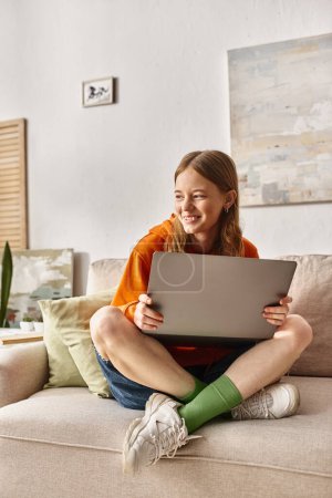 alegre adolescente con un ordenador portátil disfrutando de su tiempo y sentado en el sofá en la sala de estar