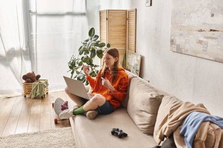 Téléchargez les photos : Adolescent heureux fille souriant lors d'un appel vidéo sur un ordinateur portable et assis près joystick sur le canapé - en image libre de droit