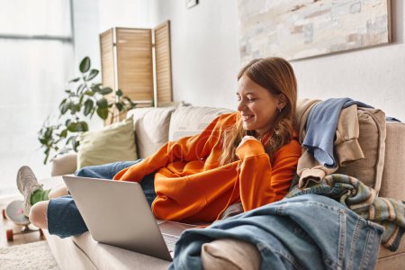 Téléchargez les photos : Heureuse adolescente regardant un film sur un ordinateur portable tout en se relaxant sur un canapé dans le salon, loisirs - en image libre de droit