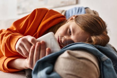 Unruhiges Teenager-Mädchen benutzt ihr Smartphone und fühlt sich im Wohnzimmer auf dem Sofa liegend