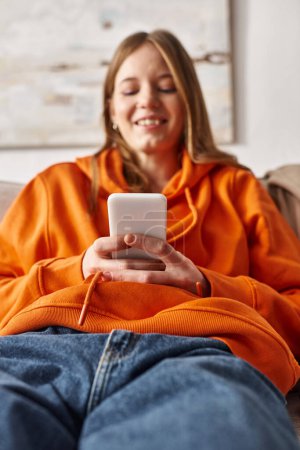 Foto de Adolescente feliz usando su teléfono inteligente y sentado en el sofá en la sala de estar, usuario de redes sociales - Imagen libre de derechos