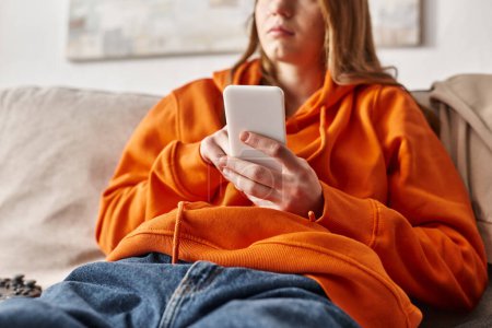 Foto de Recortado disparo de adolescente utilizando su teléfono inteligente y sentado en el sofá en la sala de estar, medios de comunicación social - Imagen libre de derechos