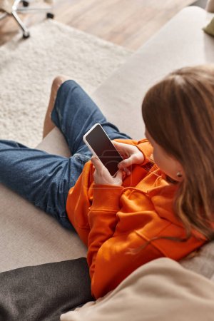 Foto de Zoomer chica adolescente usando su teléfono inteligente y sentado en el sofá en la sala de estar, usuario de redes sociales - Imagen libre de derechos