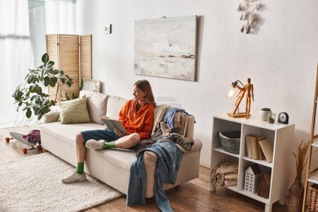 glückliches Teenager-Mädchen liest Buch, während es auf dem Sofa neben chaotischem Kleiderstapel in der Wohnung sitzt