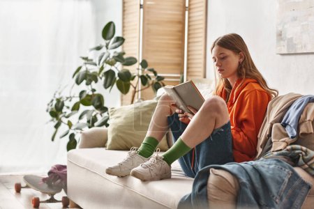 Teenagermädchen liest Buch und sitzt auf Sofa neben chaotischem Kleiderstapel in Wohnung