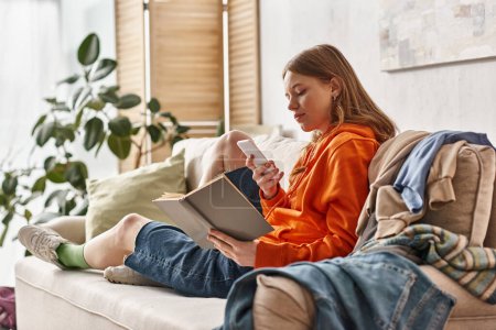 Teenager-Mädchen mit Buch und Smartphone auf Sofa neben chaotischem Kleiderstapel