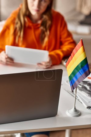 focus of lgbtq pride flag and laptop on desk near blurred teenage girl studying from home