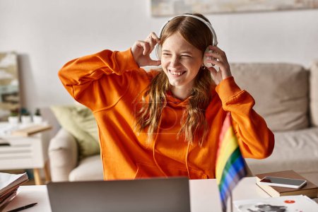 Joyful teenage girl in headphones enjoying e-learning next to laptop and pride flag on desk