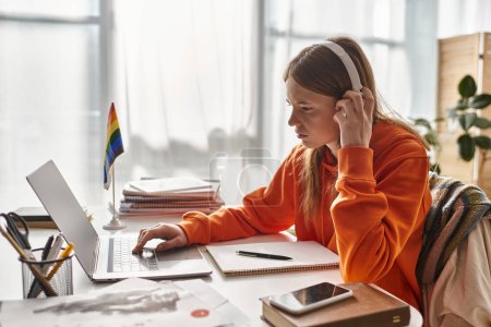 Concentrated teenage girl in wireless headphones e-learning beside pride flag and stationery