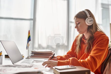Téléchargez les photos : Jeune adolescente concentrée dans les écouteurs sans fil e-learning à côté du drapeau de fierté et de papeterie - en image libre de droit