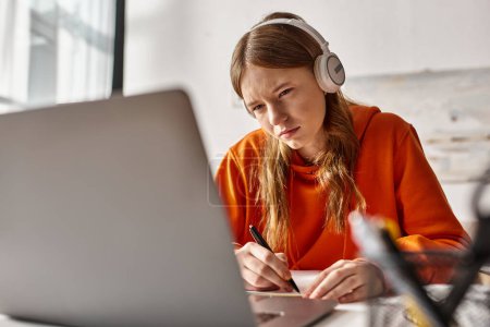 Téléchargez les photos : Jeune adolescente concentrée dans les écouteurs sans fil e-learning à côté des ordinateurs portables et de la papeterie - en image libre de droit